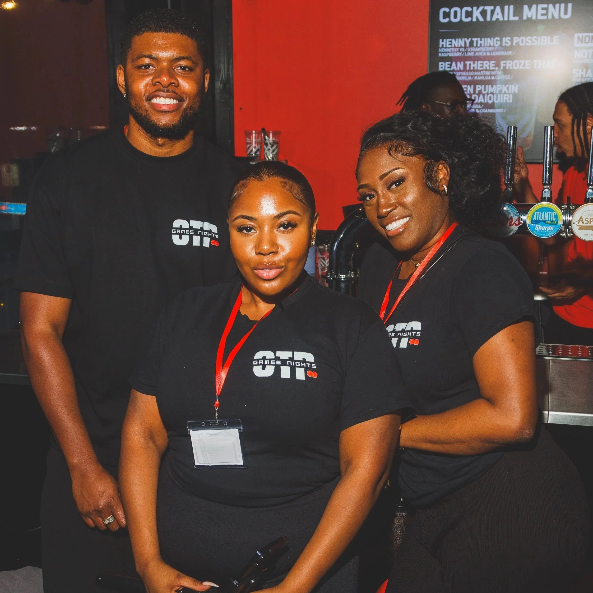 Three smiling people wearing black t-shirts with a logo reading 'OTG Games Nights' at a bar or club, with drinks and a cocktail menu in the background.