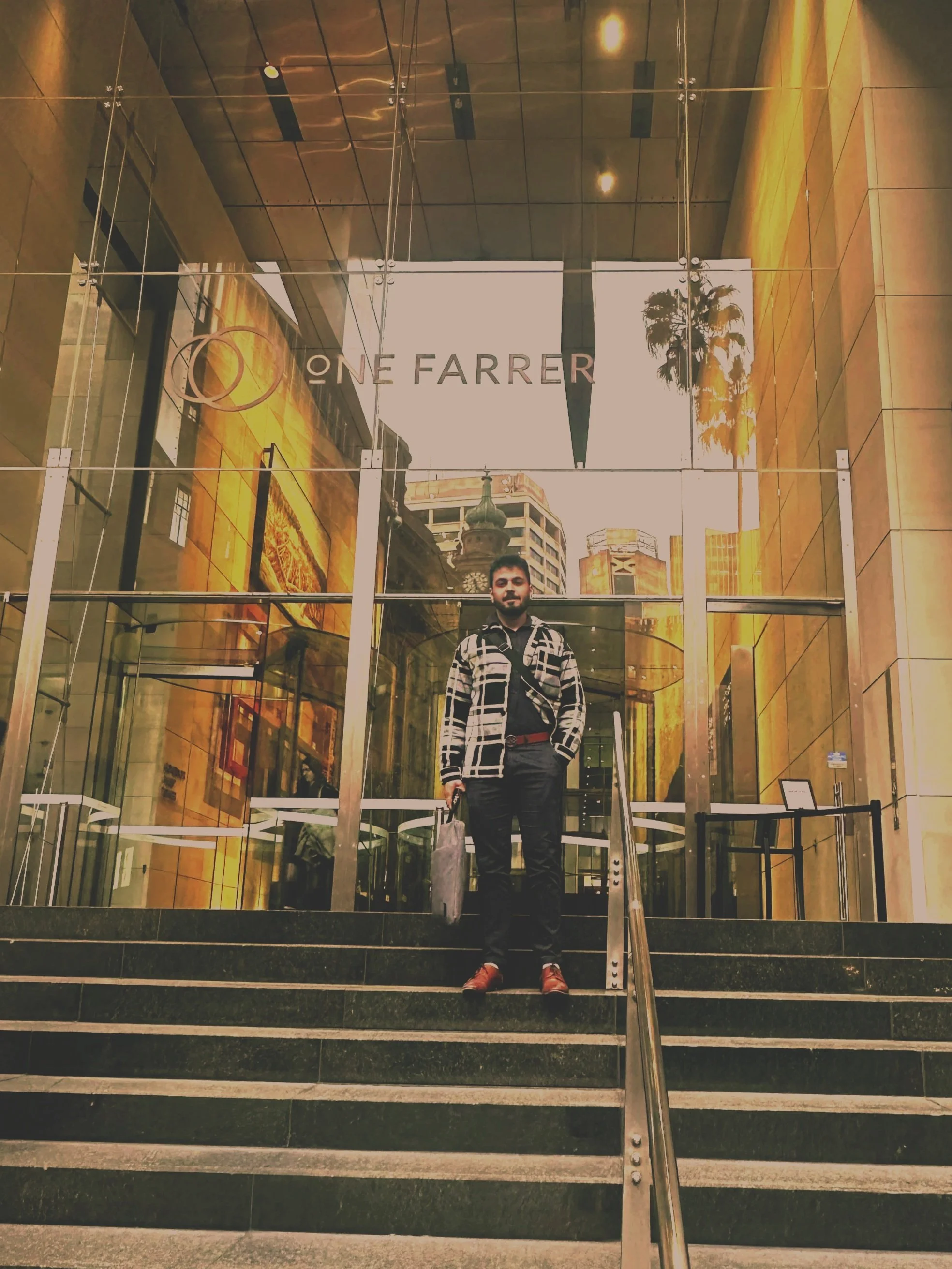 A man standing on steps outside a building with a glass entrance, the building's reflection shows tall city buildings and a palm tree. The sign on the building reads "ONE FARRER." The man is holding a bag in one hand and is dressed in a patterned jacket, black shirt, black pants, and brown shoes.