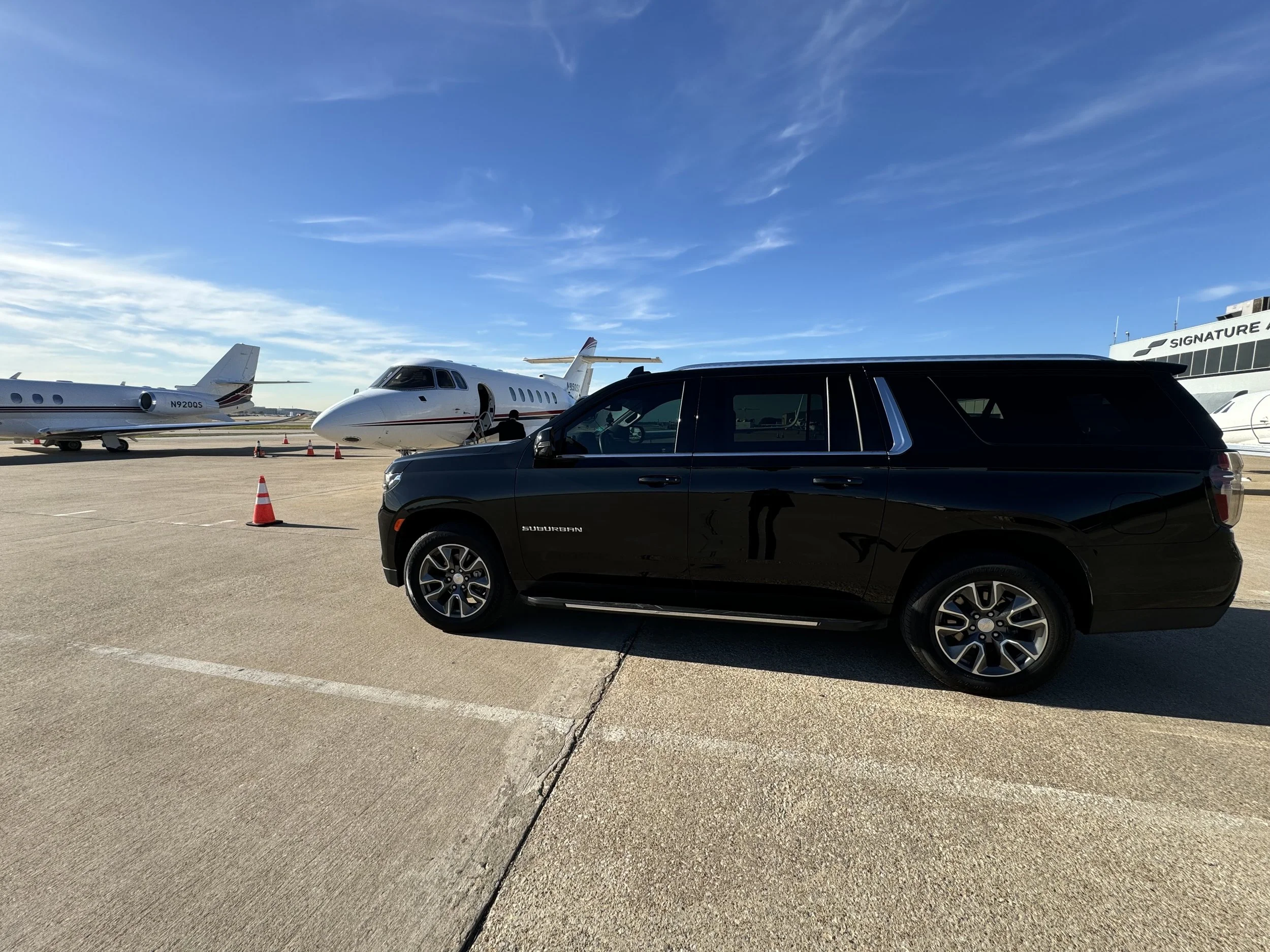 Black SUV parked on tarmac near two private jets at the Dallas Fortworth airport, with cones marking the area, under a clear blue sky.
