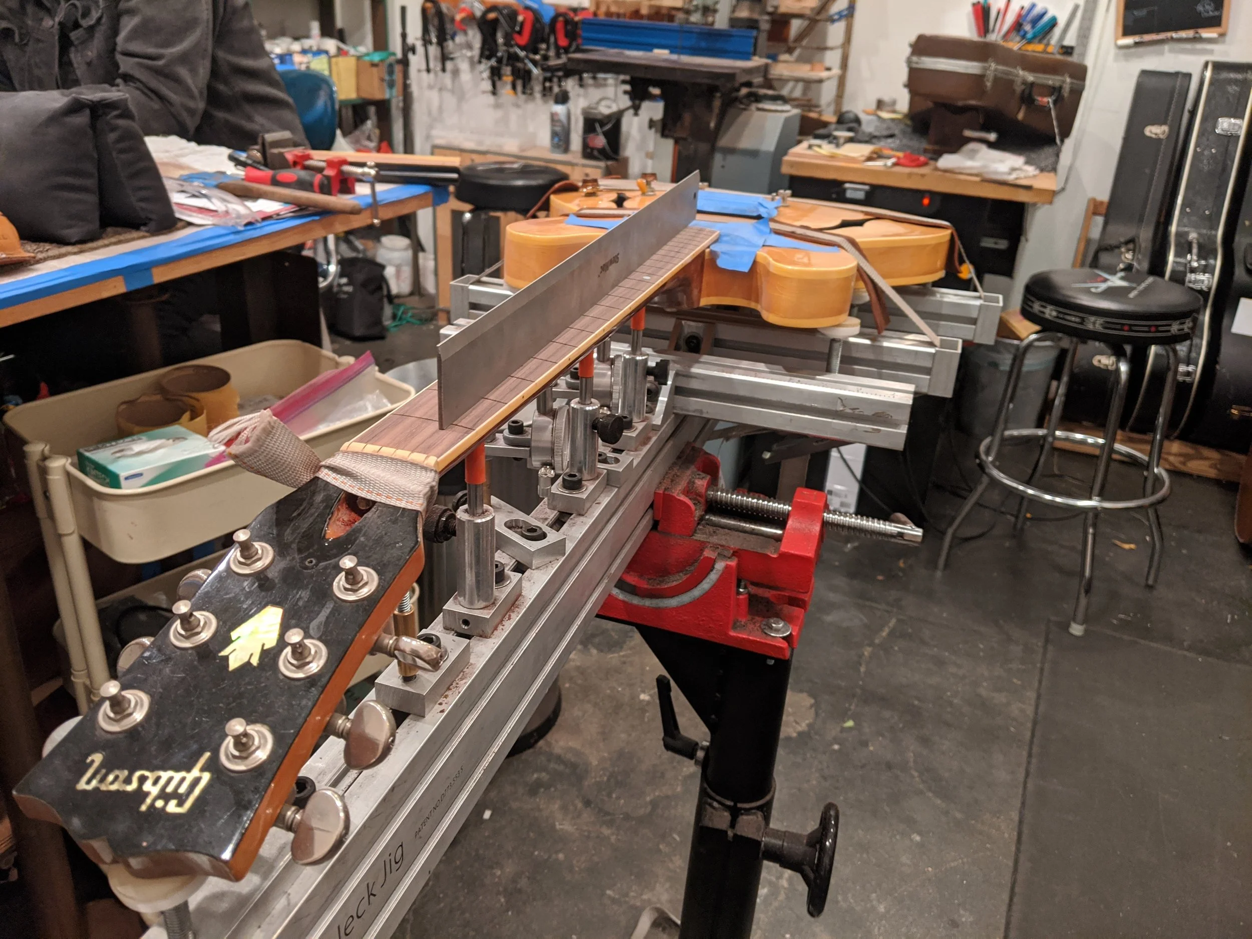 A guitar is being built on a workbench, with parts held in place by clamps, and various tools and supplies scattered around a workshop.