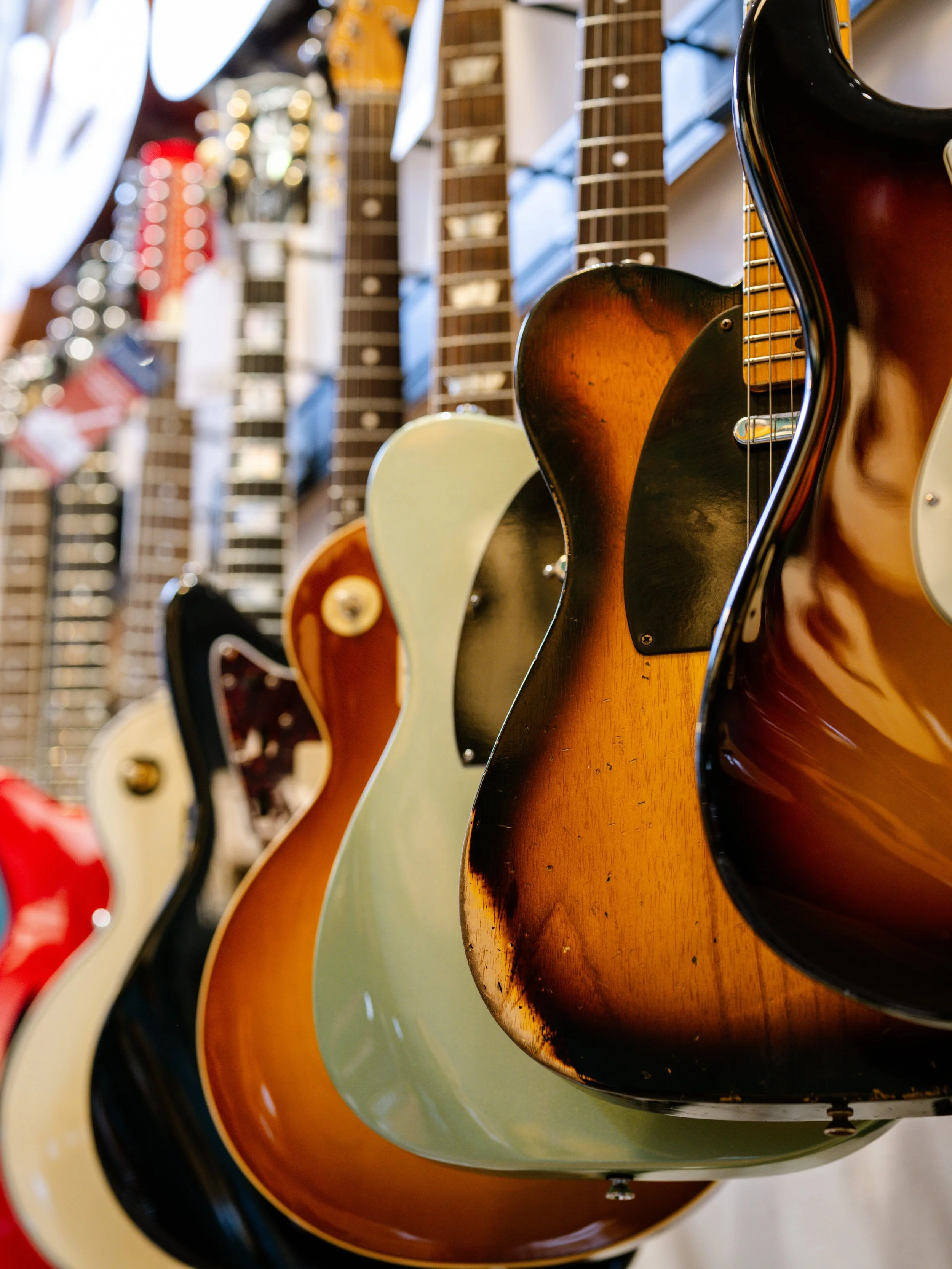 Multiple vintage guitars hanging on a wall in a music store, with various colors and styles, some with wood finishes and others with painted bodies.