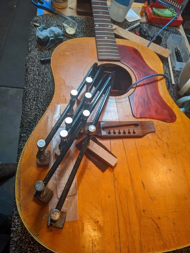 A guitar body is in the process of being repaired or assembled, with clamps securing a piece of wood to the top. The guitar is on a workbench with various tools, glue, and supplies around it.