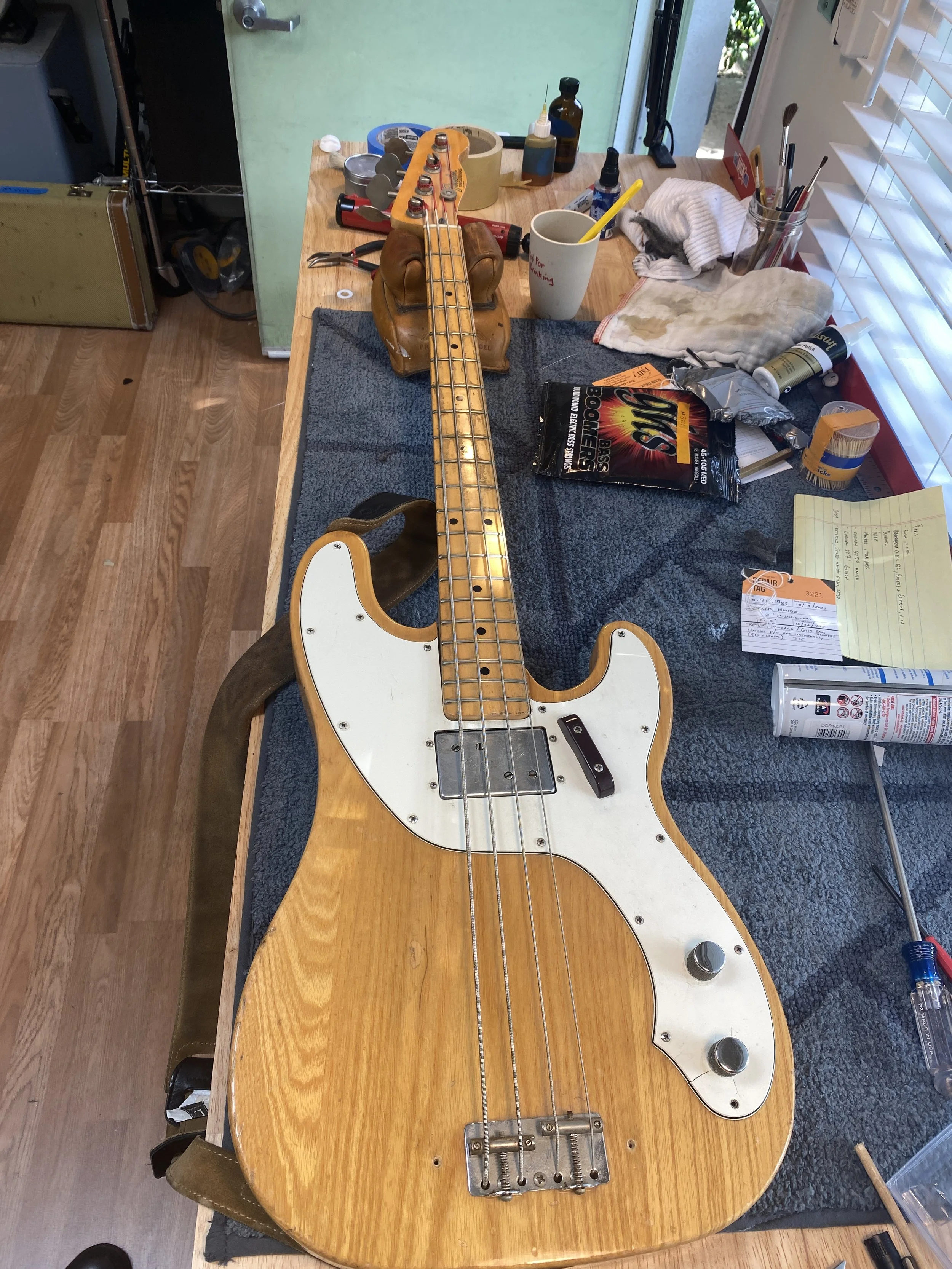 A vintage electric bass guitar with a natural wood finish and a white pickguard, placed on a worktable surrounded by tools and supplies.