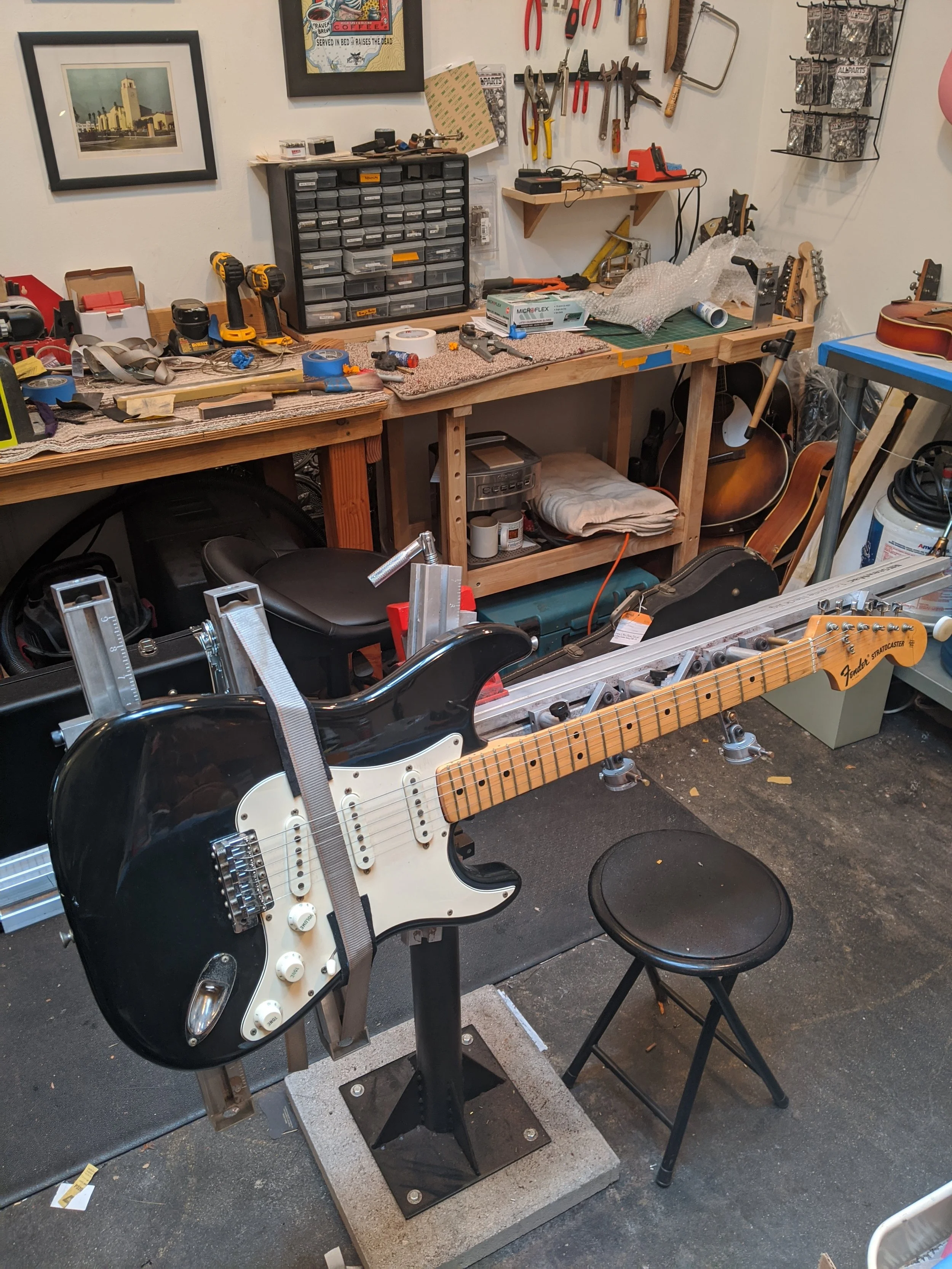A workshop with a black and white electric guitar mounted on a stand, surrounded by tools, workbenches, and chairs.