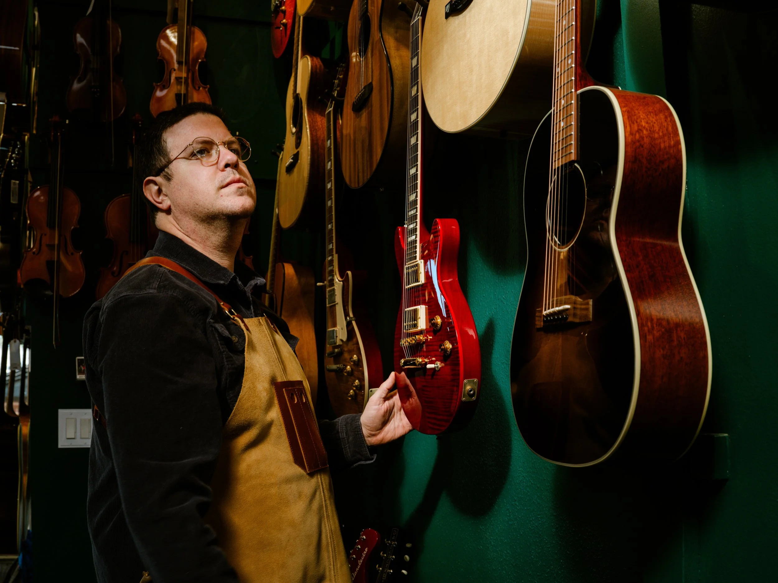 A man wearing glasses and a brown apron is examining an acoustic guitar in a music store, with other guitars hanging on the green wall behind him.