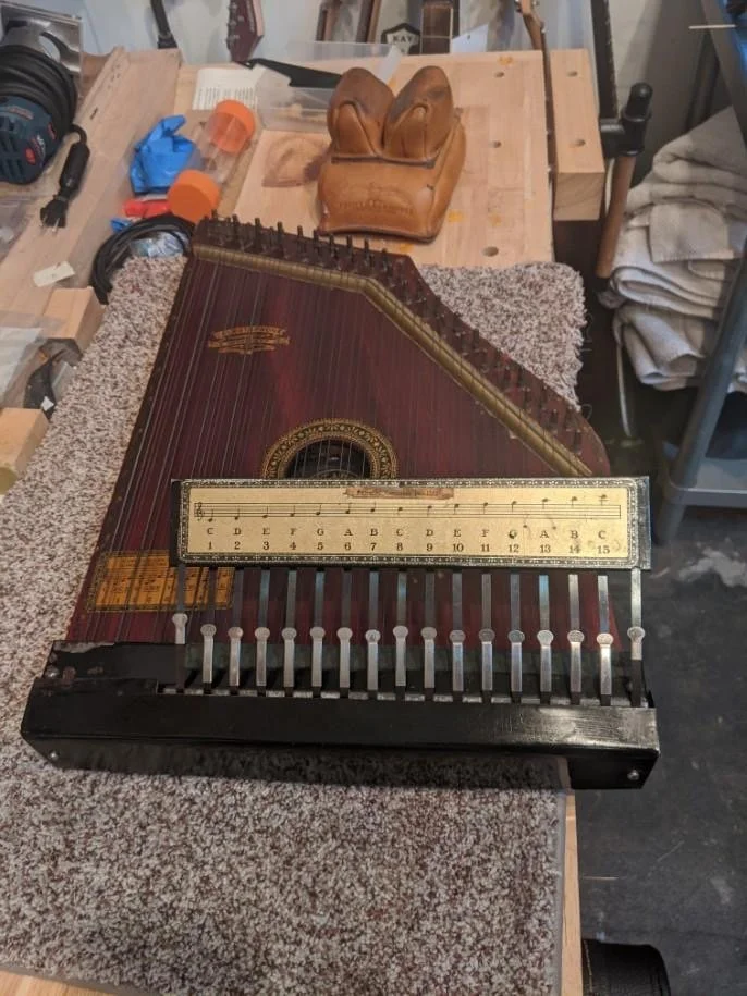 A vintage zither musical instrument with a ruler for scale, placed on a workbench in a workshop, with a leather glove, clamps, and tools in the background.