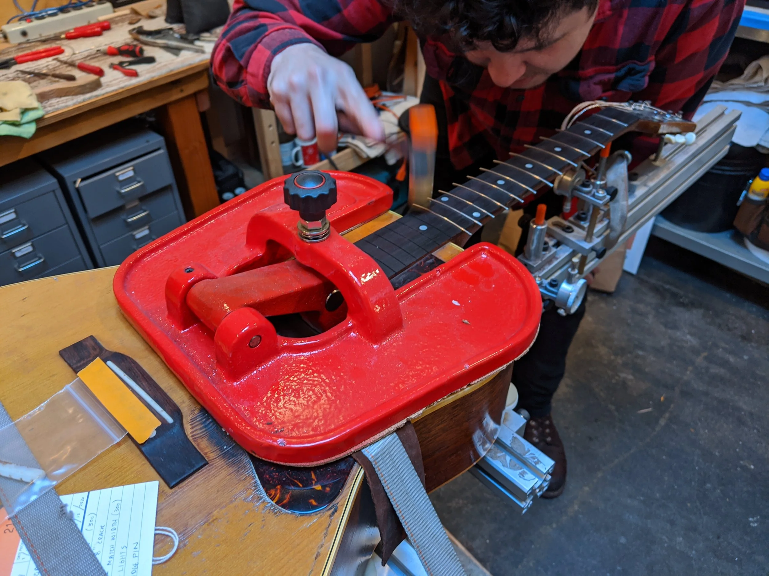 A person working on a guitar neck secured in a red woodworking jig on a workbench in a workshop, with tools and storage drawers in the background.