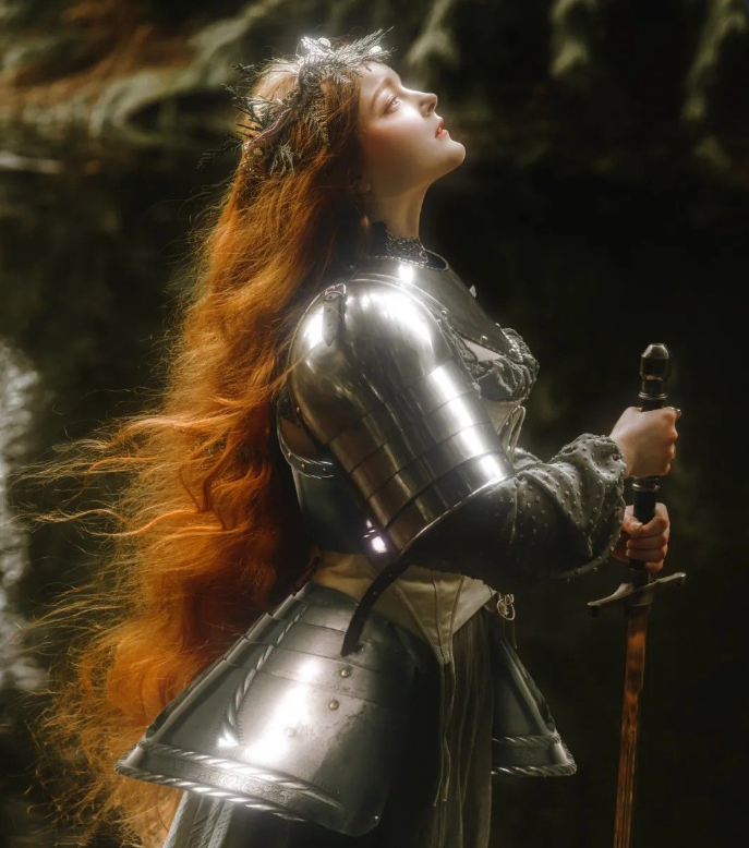 A woman dressed as a medieval warrior with long, flowing red hair, wearing shiny armor and holding a sword, standing outdoors near water.