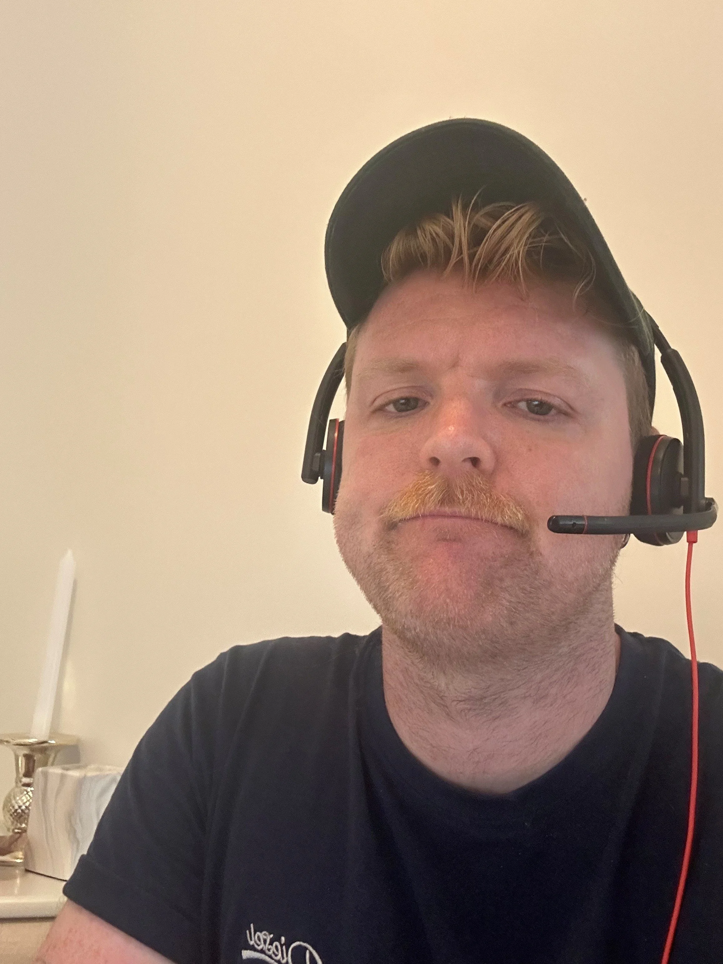 A man with a blond mustache and beard wearing a black snapback cap, black shirt, and a headset with microphone, taking a selfie indoors.