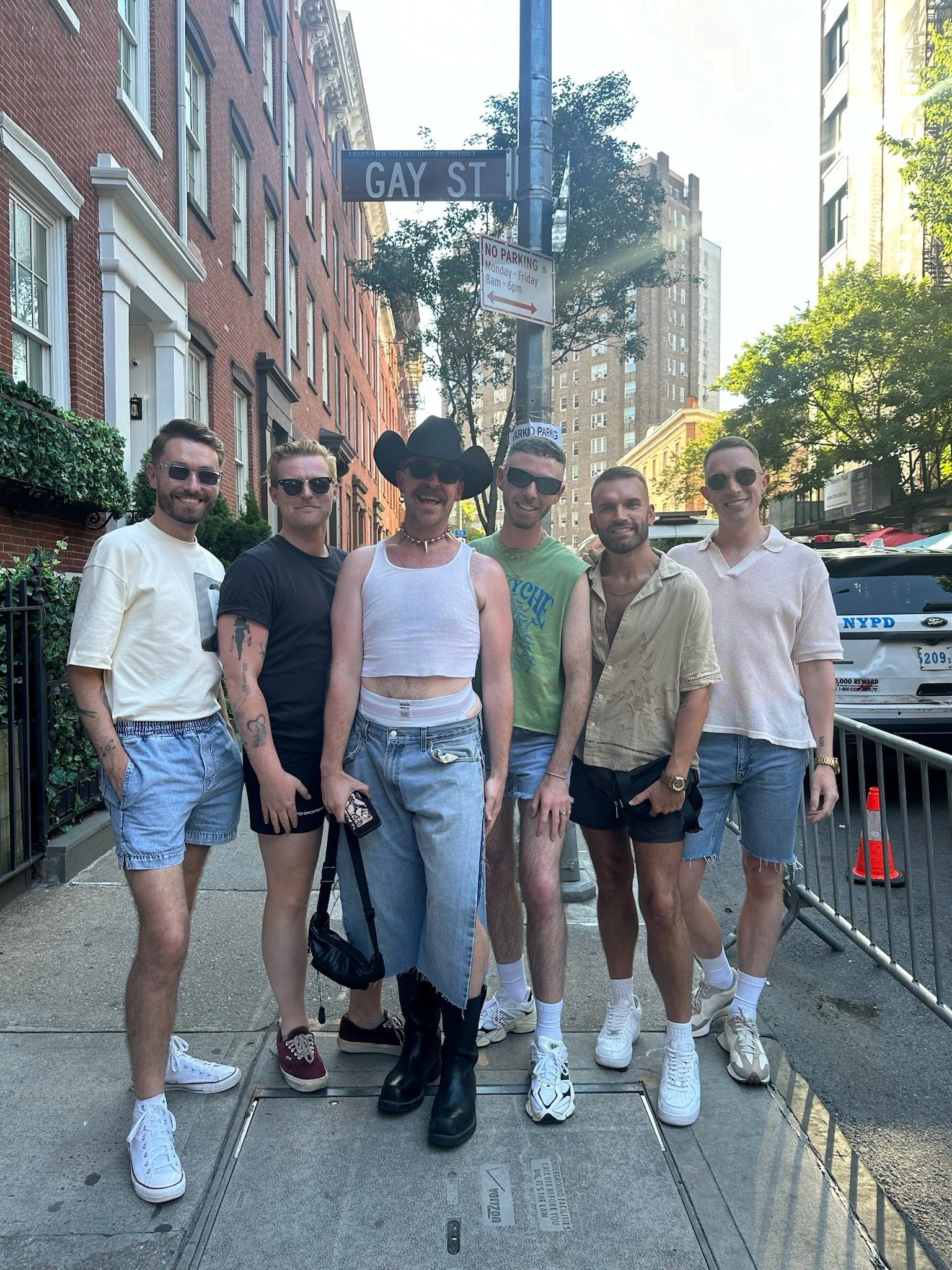 Group of seven young adults standing on a city sidewalk in front of a street sign reading "Gay St". They are wearing casual summer clothes and sunglasses, smiling at the camera. City buildings and trees are visible in the background.