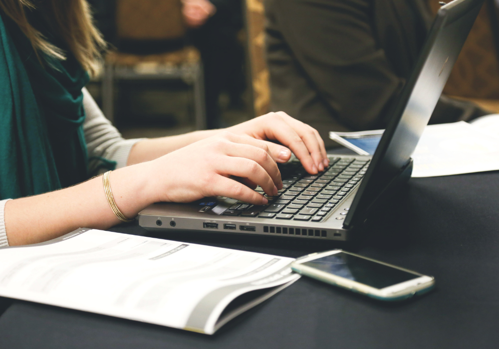 A person typing on a laptop at a desk with papers and a smartphone nearby.