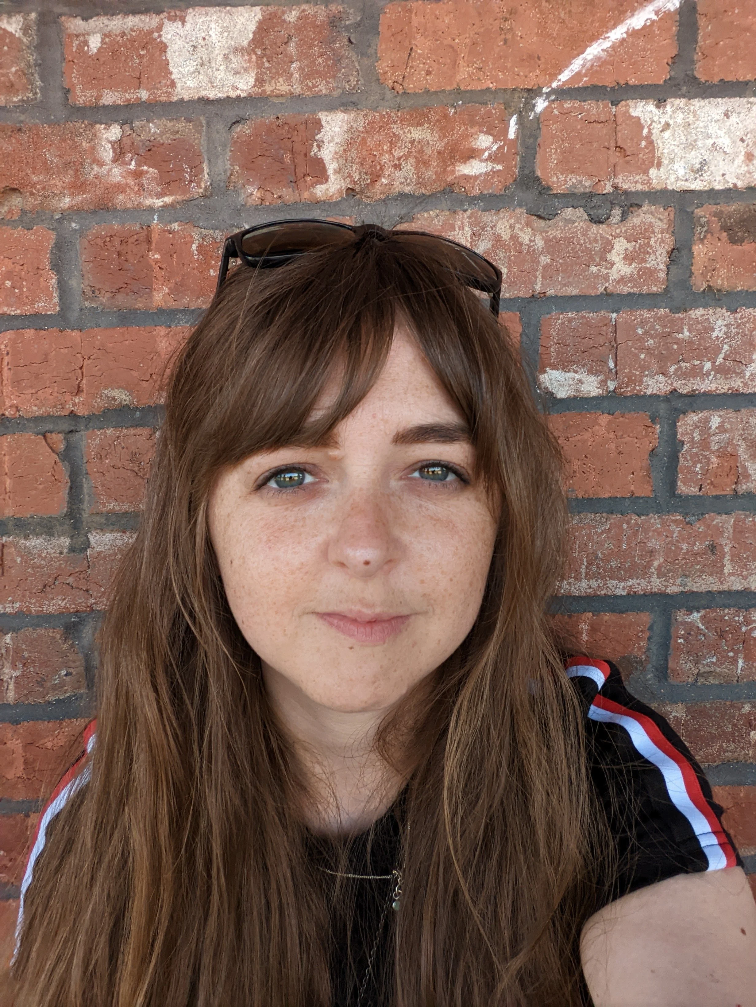 A young woman with long, reddish-brown hair, light blue eyes, and fair skin with freckles, wearing sunglasses on her head and a black shirt with red and white stripes on the sleeves, standing in front of a brick wall.