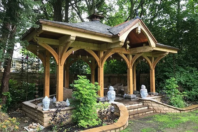 Wooden gazebo in a backyard with stone steps, surrounded by greenery and garden statues.