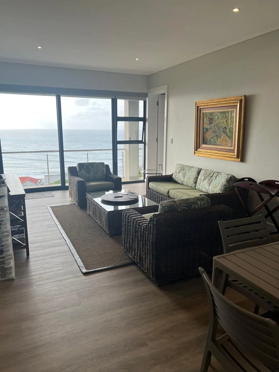 Living room with ocean view, wicker sofa and chairs, glass coffee table, framed artwork, sliding glass doors, and wooden dining table with chairs.