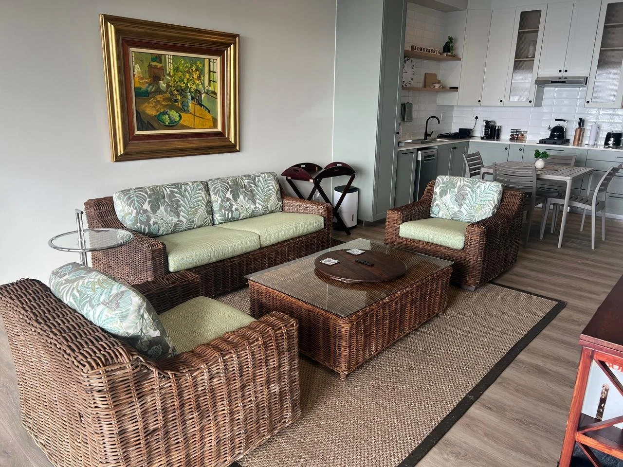 Living room with wicker furniture, a glass-topped coffee table, and a framed painting on the wall, adjacent to an open kitchen and dining area.