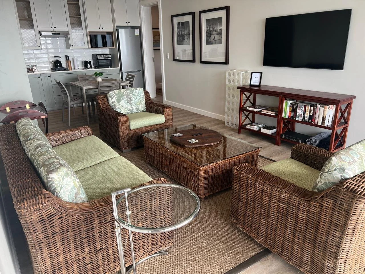 Living room with wicker furniture, a glass-top coffee table, a flat-screen TV, a bookshelf, and a dining area with a table and chairs.