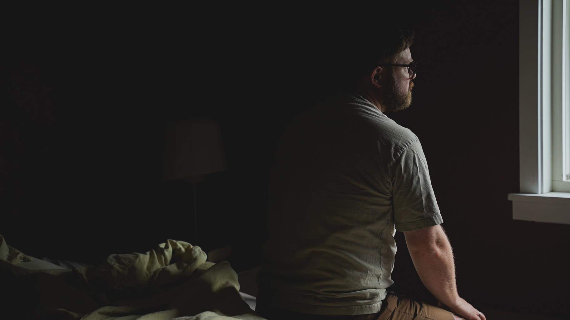 A man sitting in a half lit room, staring thoughfully out the window.