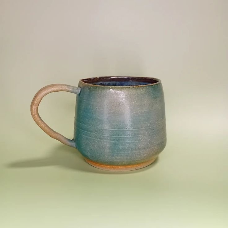 A ceramic mug with a blue-green glaze and a tan handle, placed on a light green surface with a plain light background.