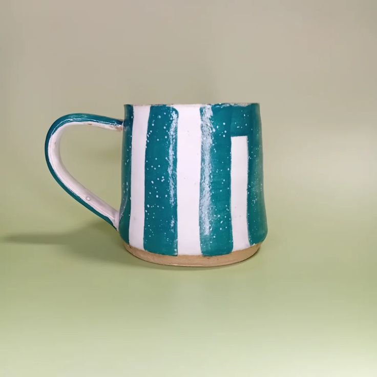 Handmade ceramic mug with white and teal vertical stripes on a light green background.