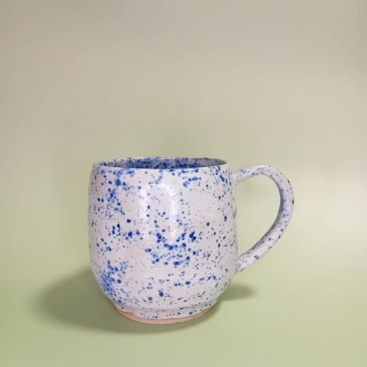 A speckled ceramic mug with a white background and blue dots, placed on a light green surface against a light green wall.
