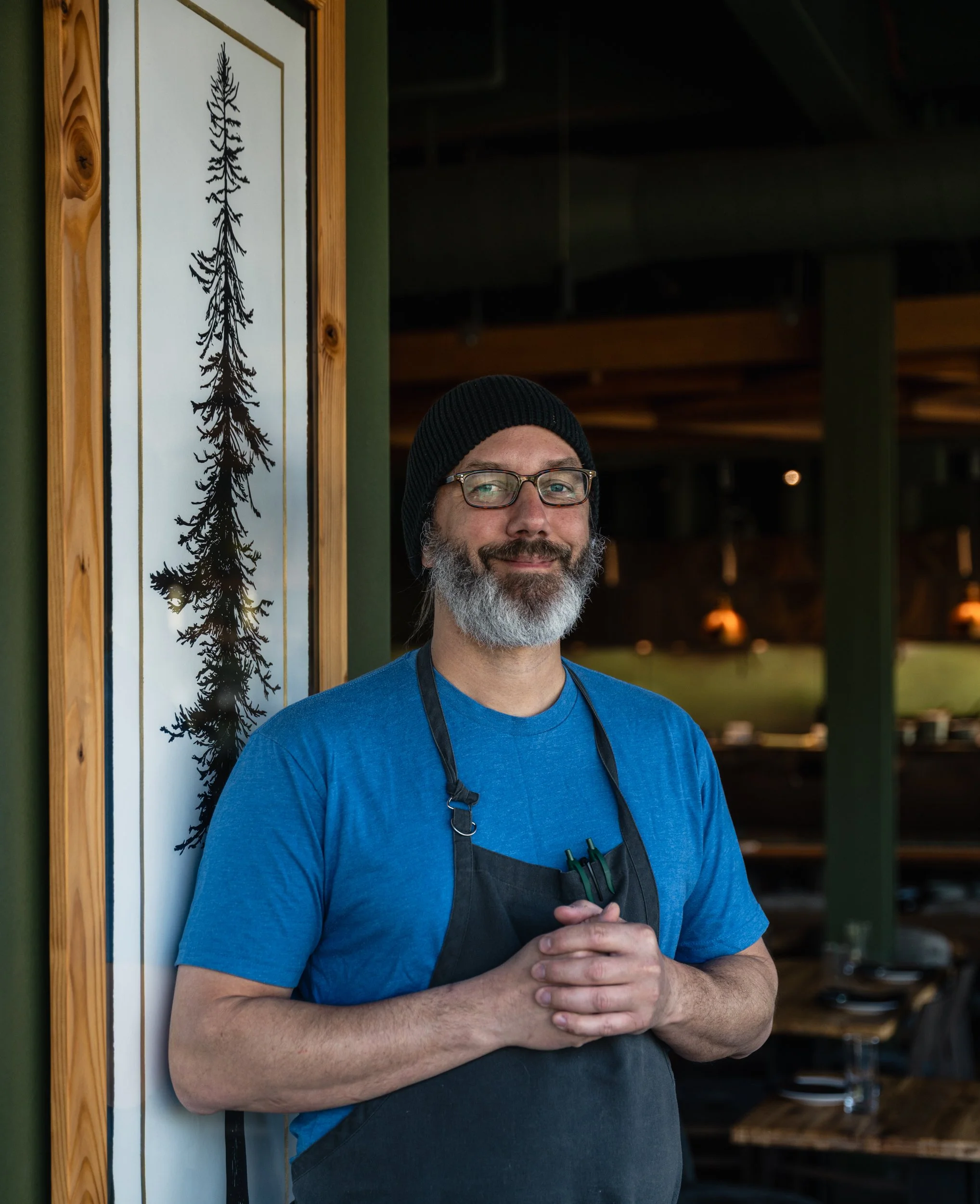 A man with a beard and gray hair smiling, wearing a black t-shirt and an apron, standing in front of a painted backdrop of trees and nature.