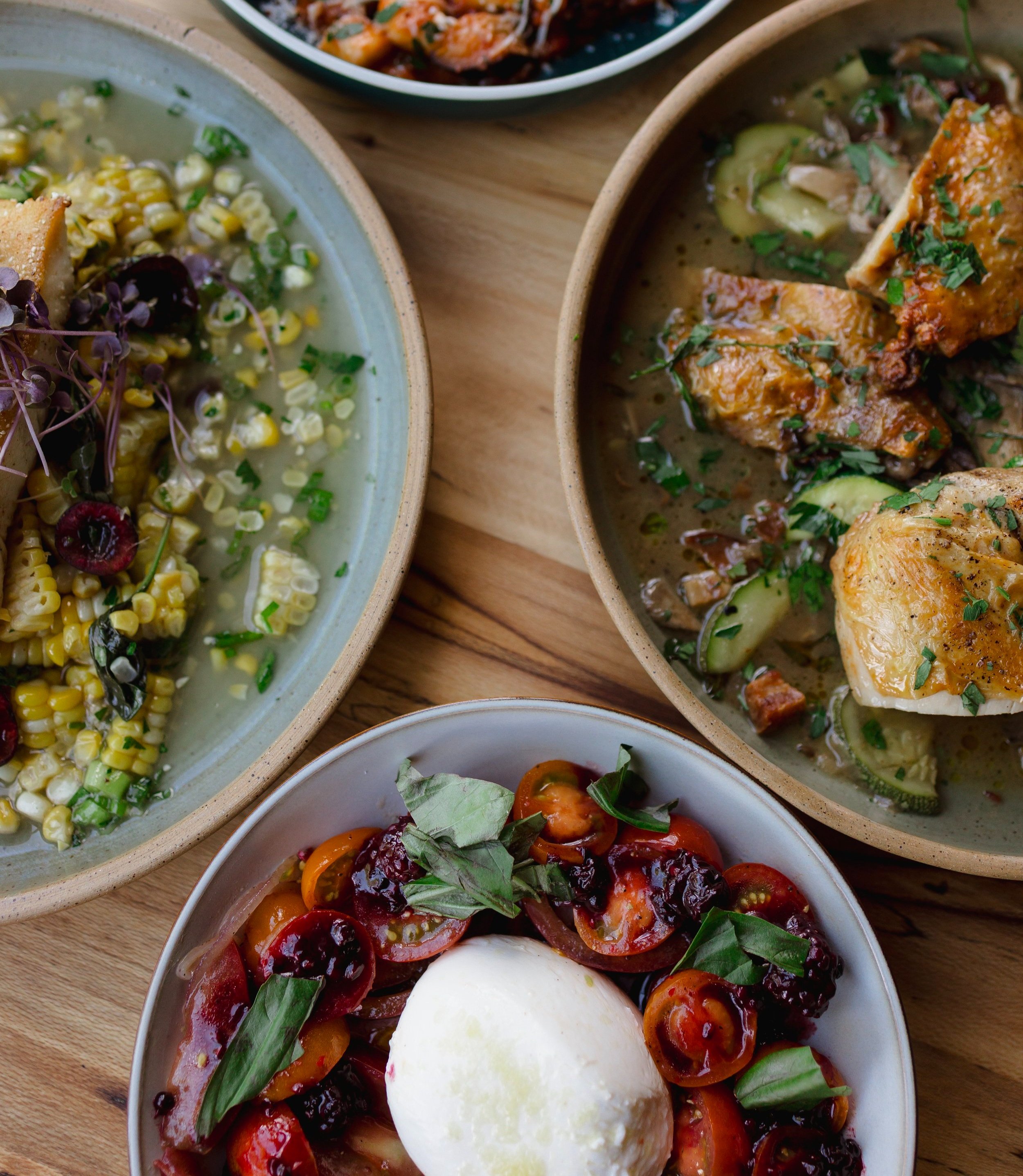 Assorted dishes including a corn salad with herbs and cherries, a chicken and zucchini stew, and a caprese salad with cherry tomatoes, basil, and a poached egg.
