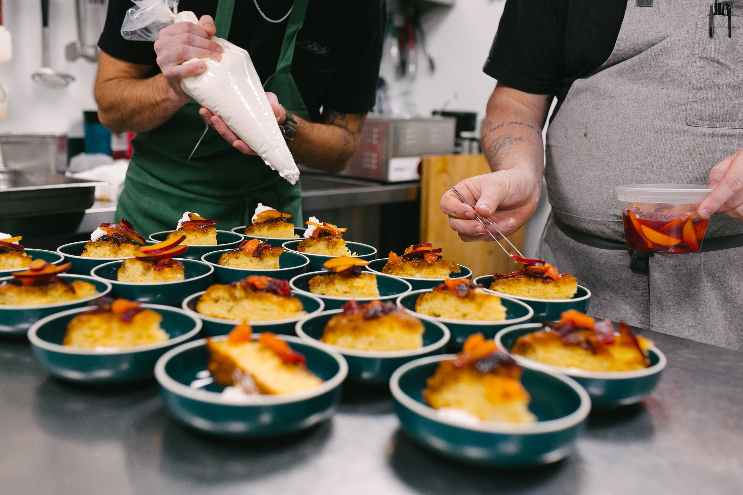 Two chefs prepare individual servings of cake with fruit toppings, using piping bags and tweezers in a professional kitchen.