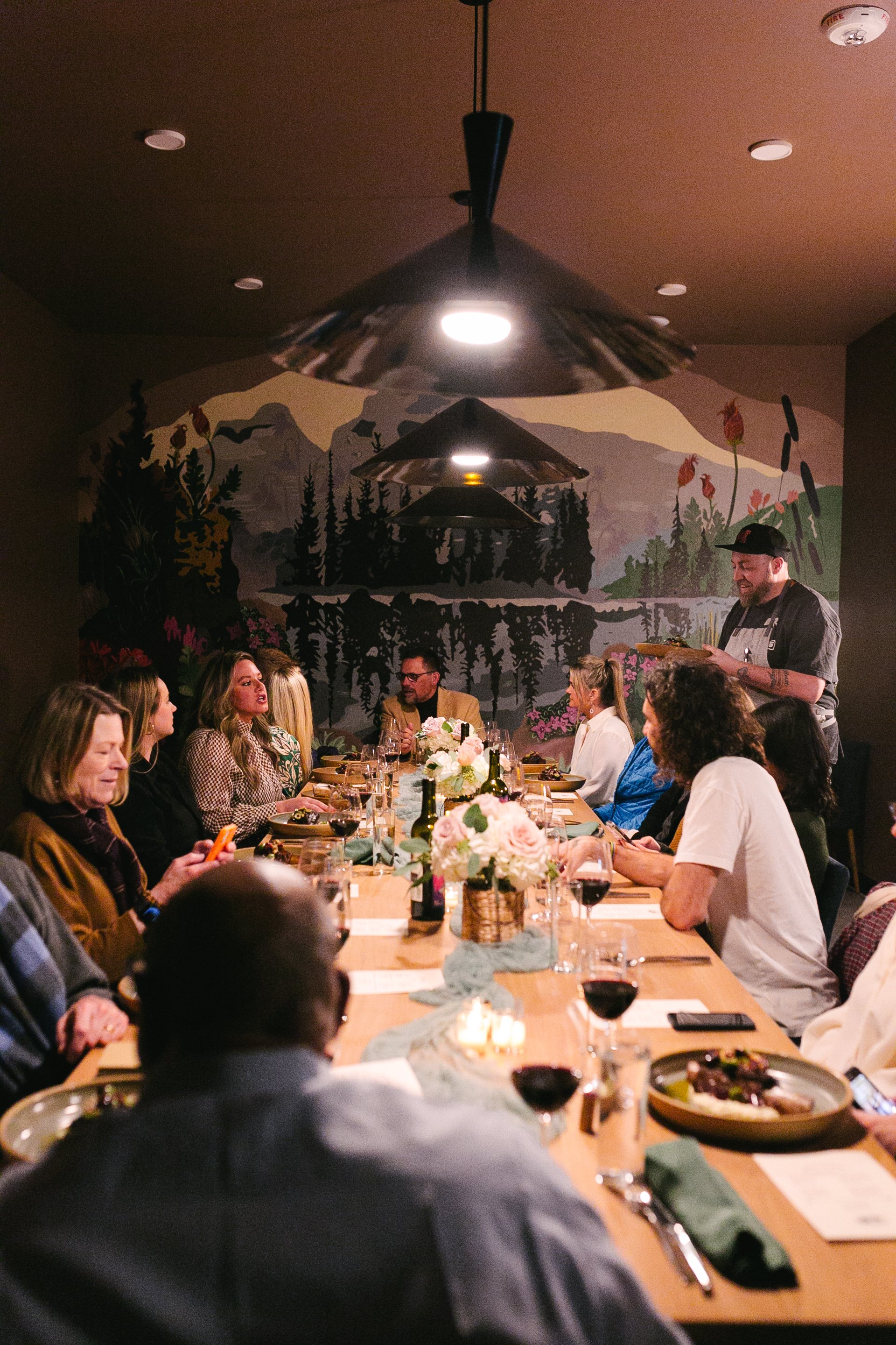 People sitting at a long dinner table in a dimly lit restaurant with floral arrangements and a mural of mountains and trees on the wall.