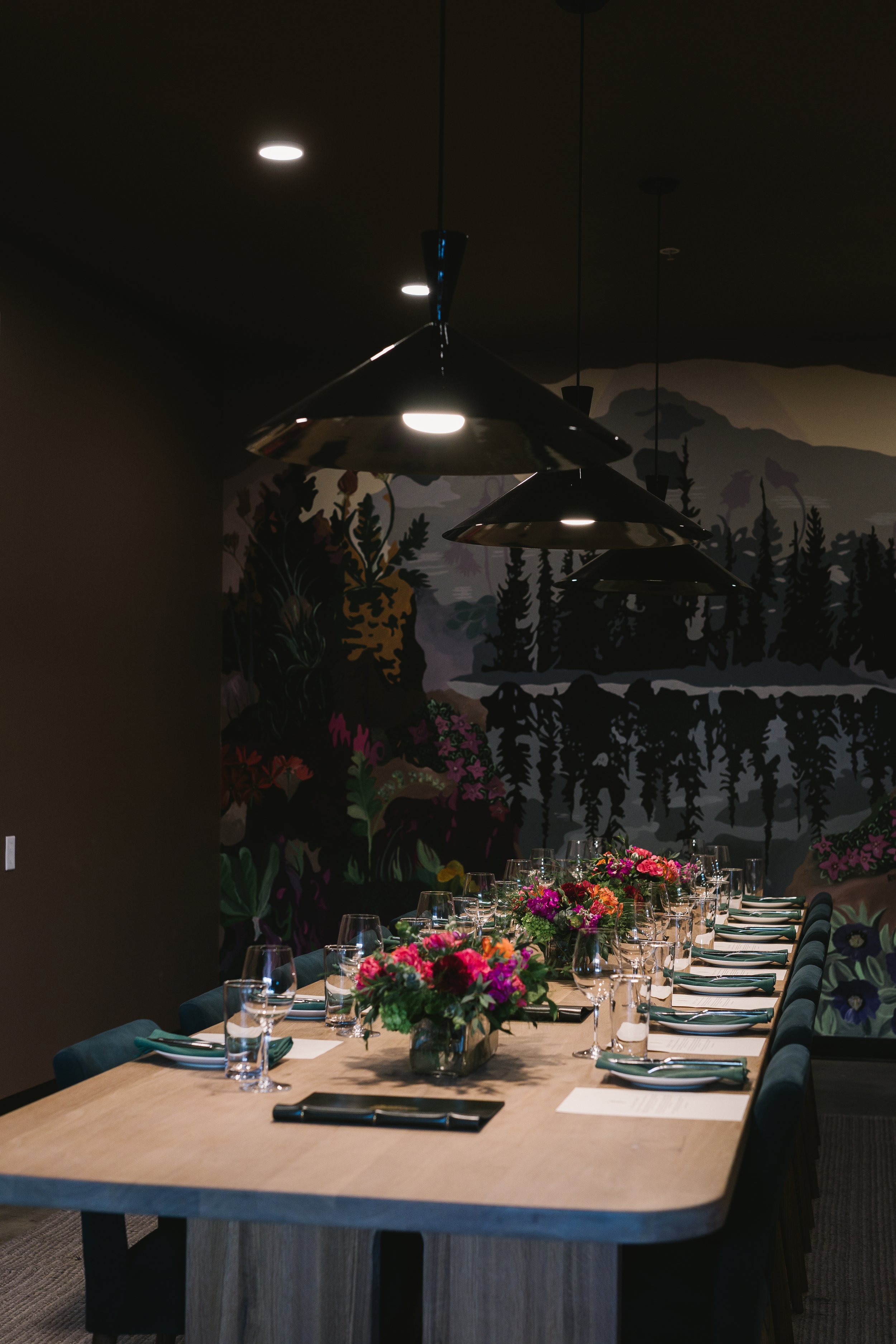 A dining room table set for a meal with floral centerpieces, wine glasses, and napkins, under three black pendant lights, with a mural of a forest scene on the dark wall in the background.