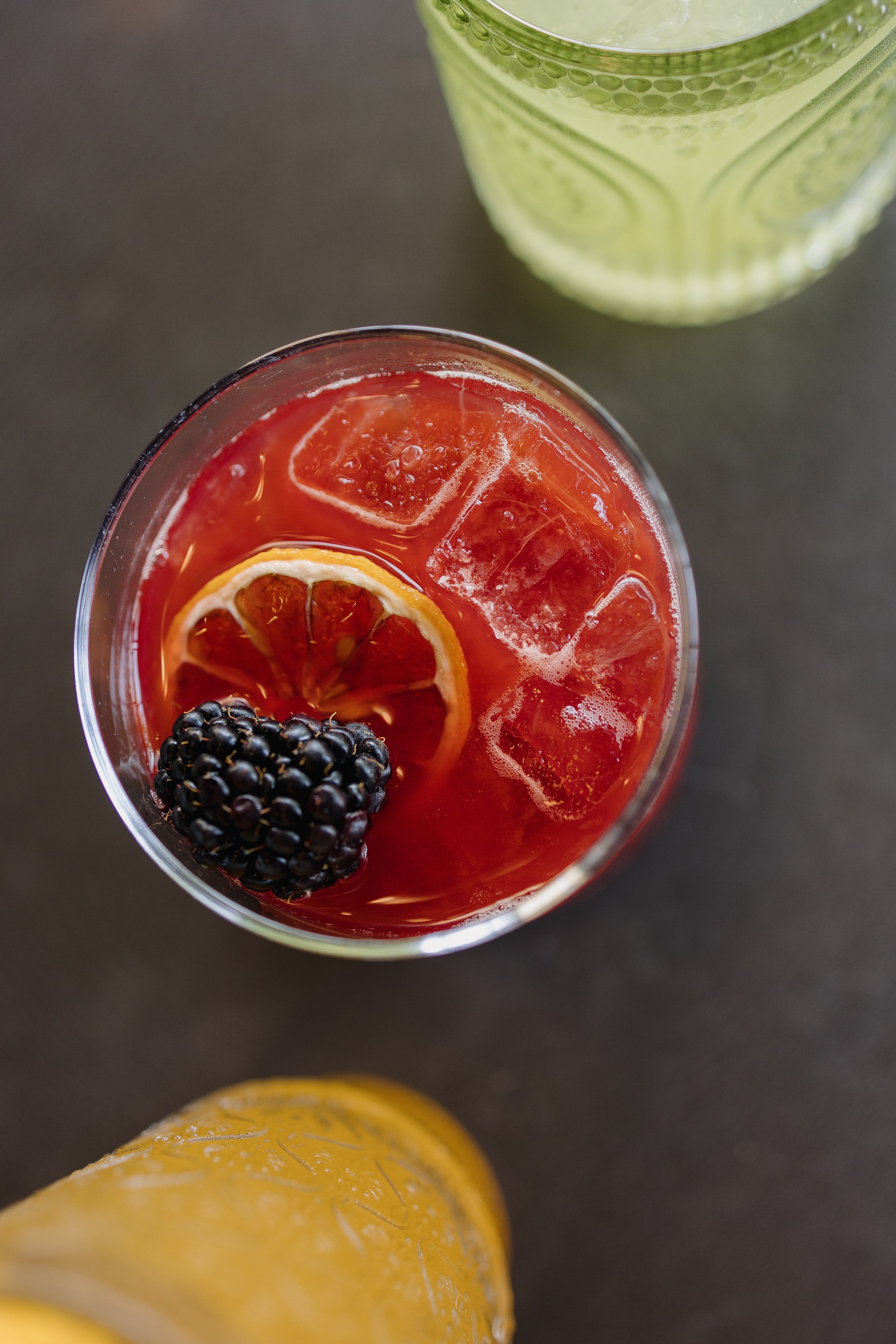 A top-down view of a glass of red fruit punch with ice, a slice of lemon, and a blackberry, with a green drink and a yellow drink partially visible in the background.