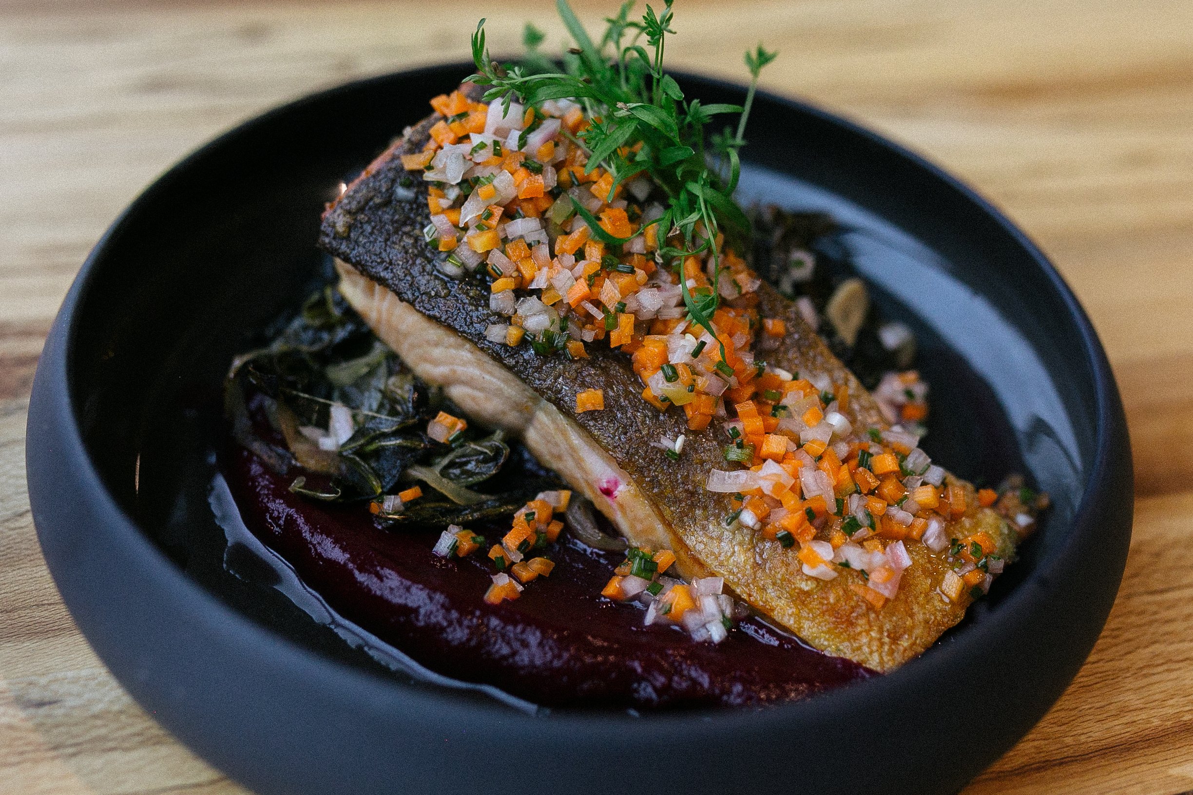 A cooked piece of fish with crispy skin topped with chopped vegetables and herbs, served on a black dish with a dark sauce underneath and a garnish of fresh herbs.