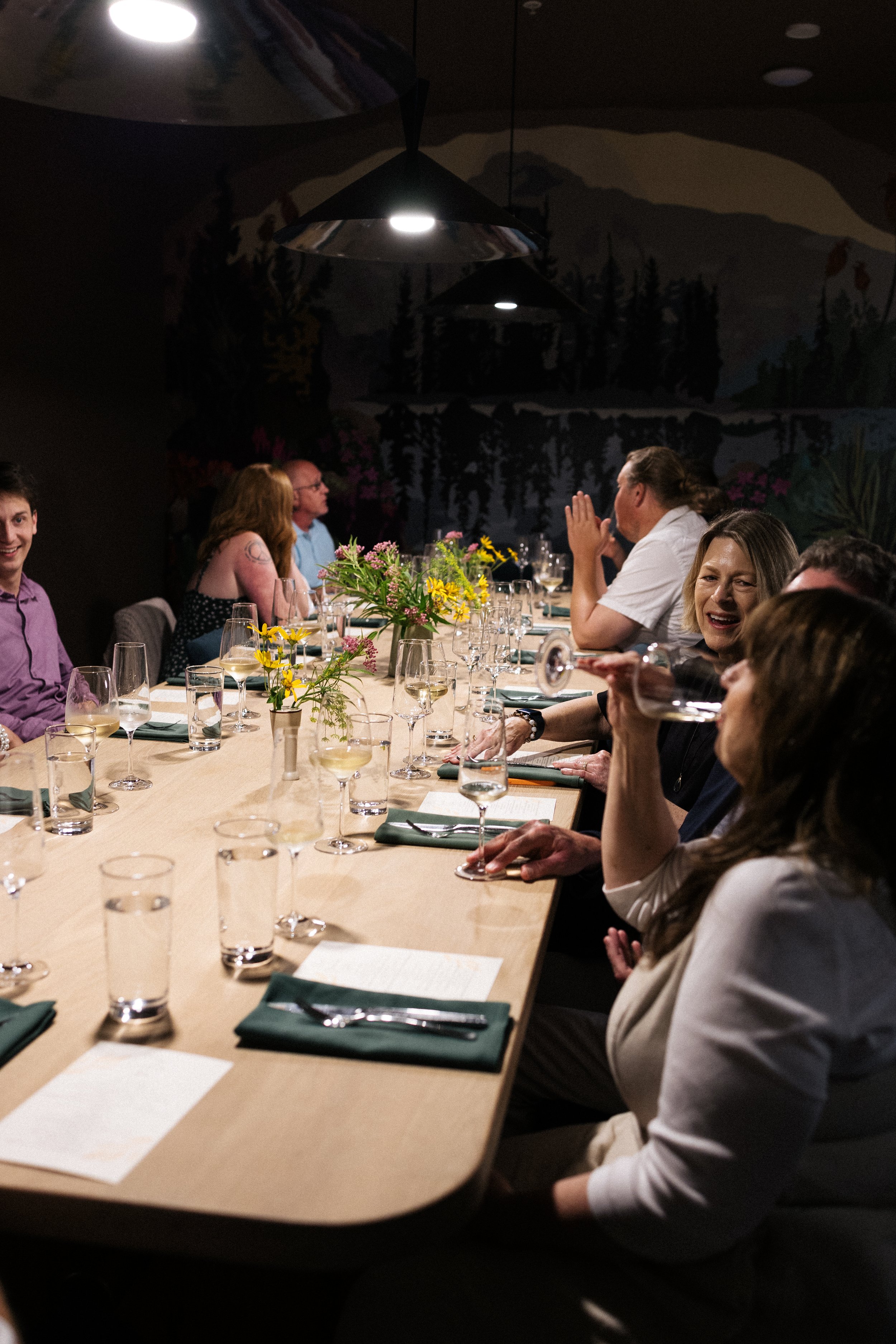 People gathered around a dining table with glasses of wine, floral centerpieces, and table settings, in a dimly lit room with a mountain mural on the wall.