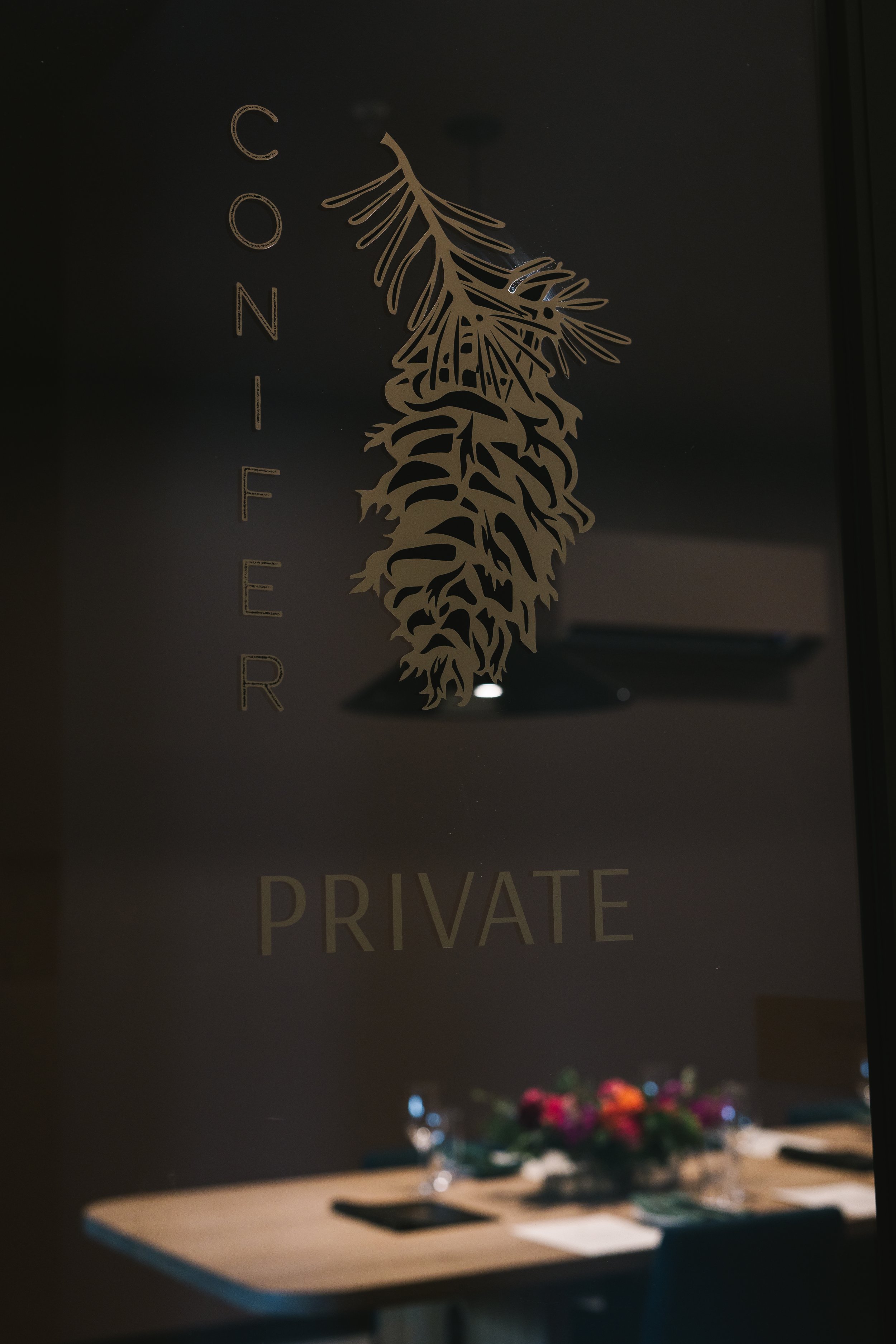 A glass wall with gold text reading "CONFER" vertically and "PRIVATE" horizontally, with a gold decorative pine cone or leaf design at the top, and a table with a floral centerpiece inside a dimly lit room.