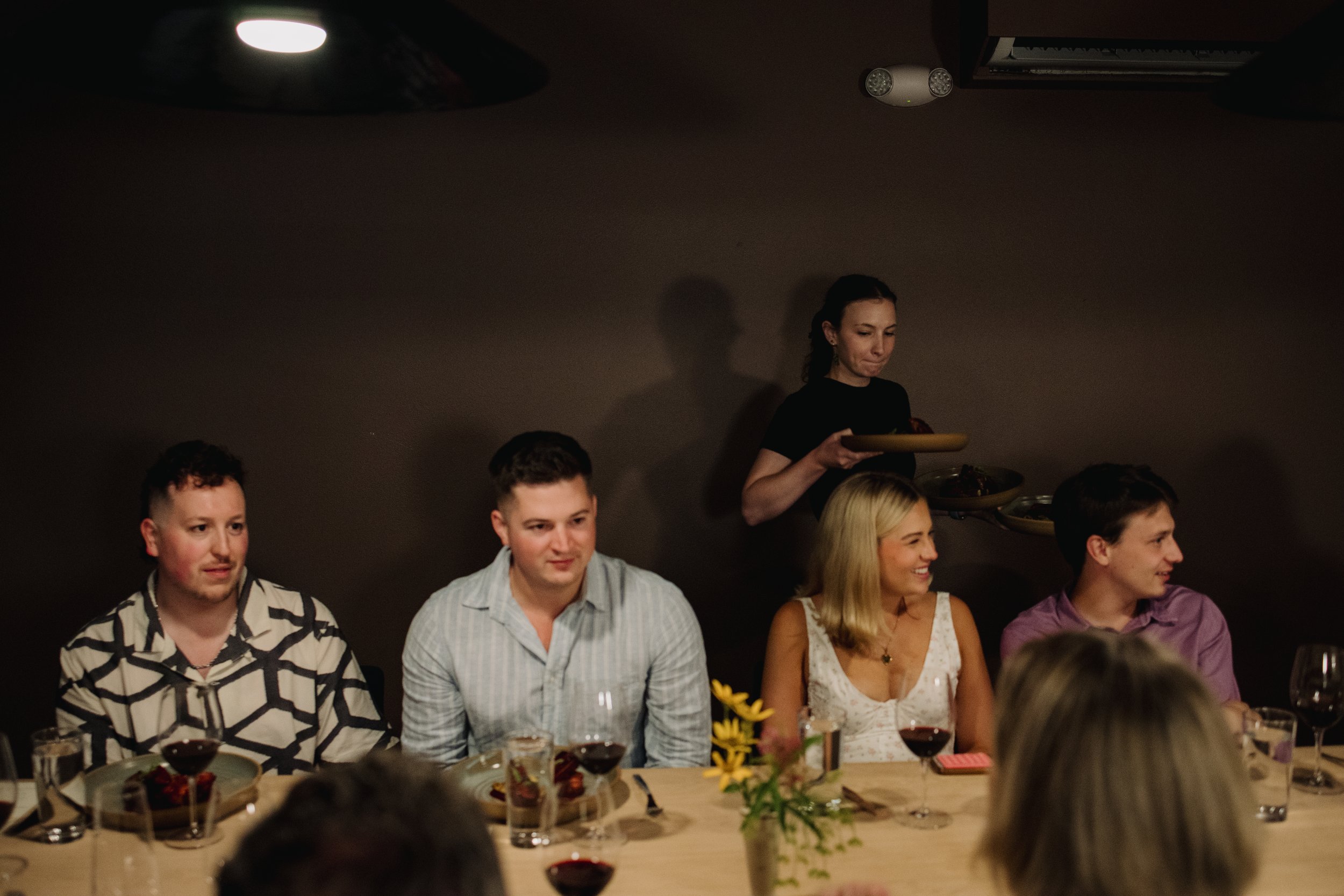 People sitting at a dinner table with wine glasses, with a woman serving food in the background.