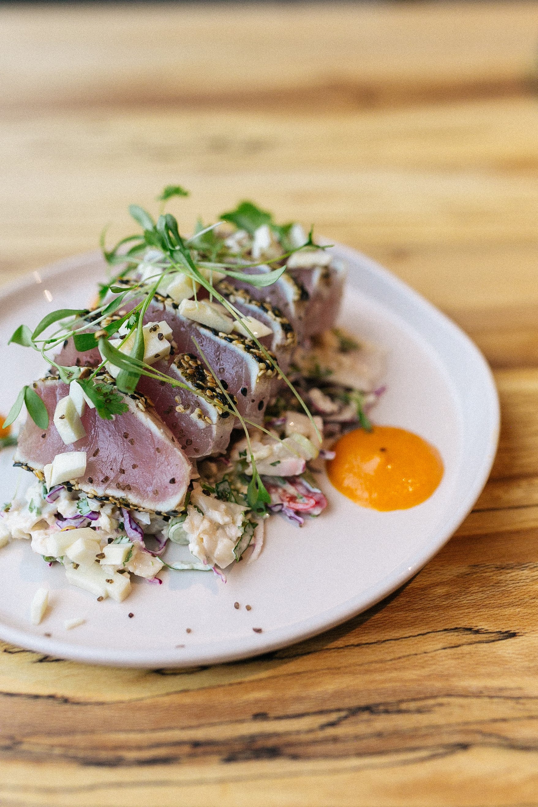 A plate of sliced seared tuna topped with microgreens, served with a side of potato salad and a dollop of orange sauce on a wooden table.