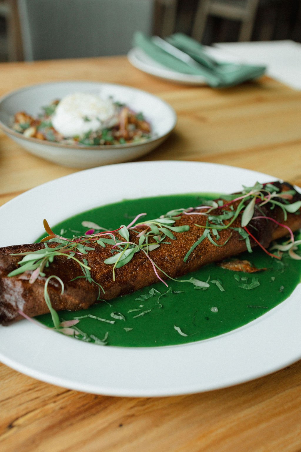 A plated dish featuring a large, crispy chimichanga garnished with microgreens on a green sauce, with a bowl of a similar dish and table settings in the background.