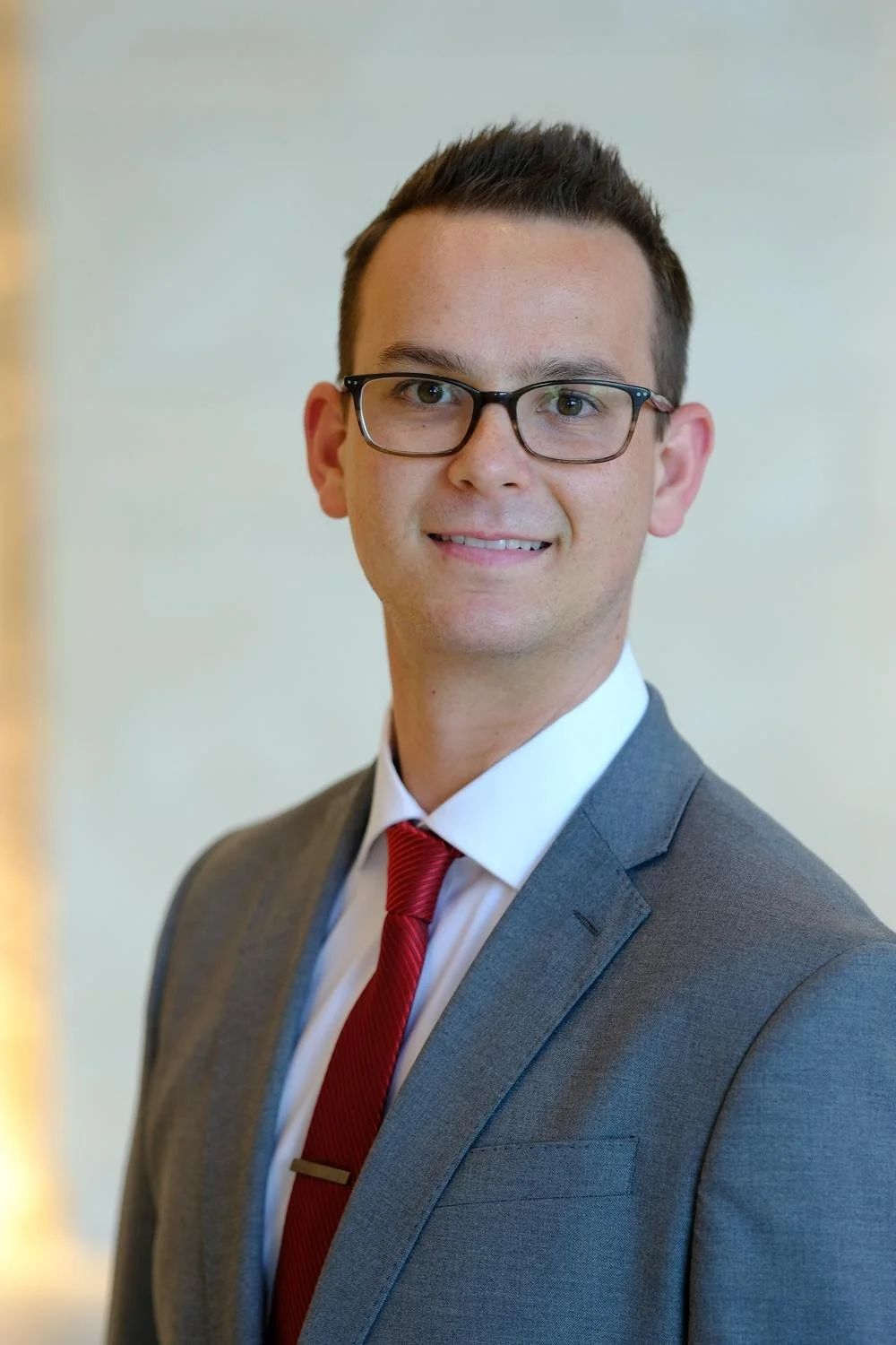 Professional headshot of a young man in a grey suit, white shirt, red tie, and glasses, smiling at the camera.