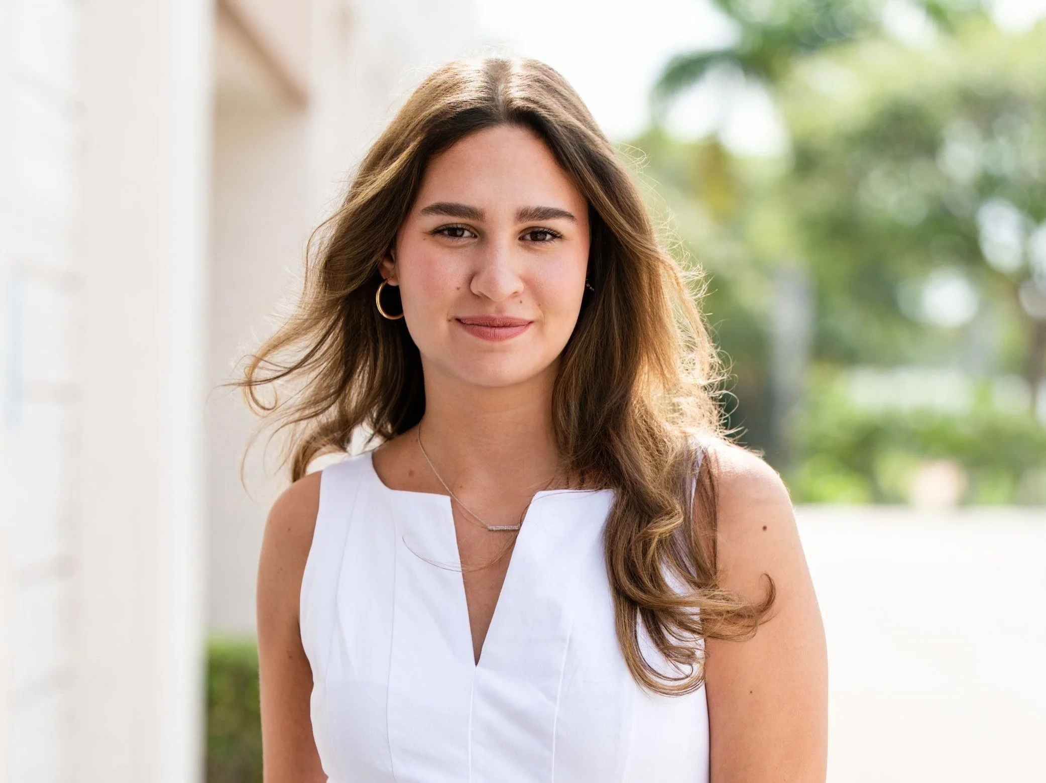 A young woman with long wavy brown hair, wearing a white sleeveless top, gold hoop earrings, and a delicate necklace, standing outdoors with a blurred green background.