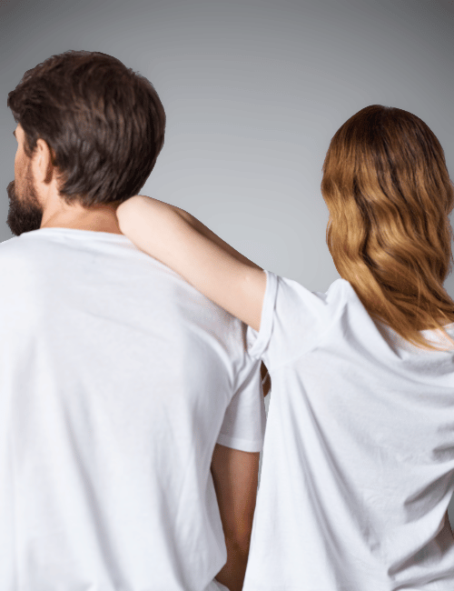 A woman with wavy red hair has her arm around the shoulder of a man with dark hair and a beard, both wearing white t-shirts.