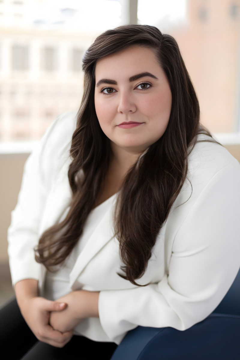 A woman with long brown hair and light skin is smiling at the camera, sitting indoors.