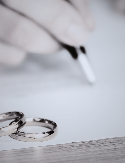 Close-up of two wedding rings on a table with a hand holding a pen in the background.