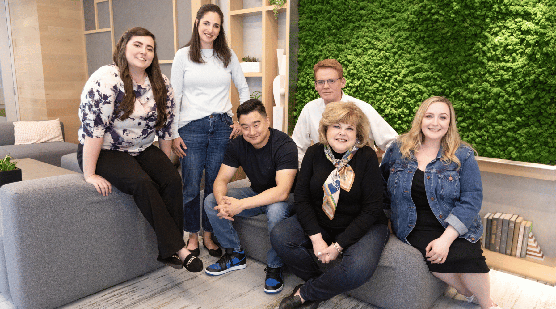 Group of seven diverse people smiling and posing together in a modern office lounge with green wall art and books.