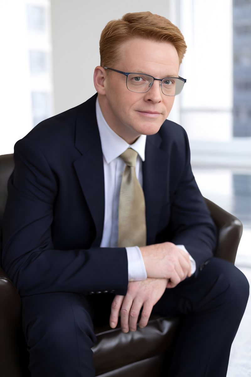 A man with red hair wearing a light gray shirt and a smartwatch sitting outdoors with green foliage in the background.