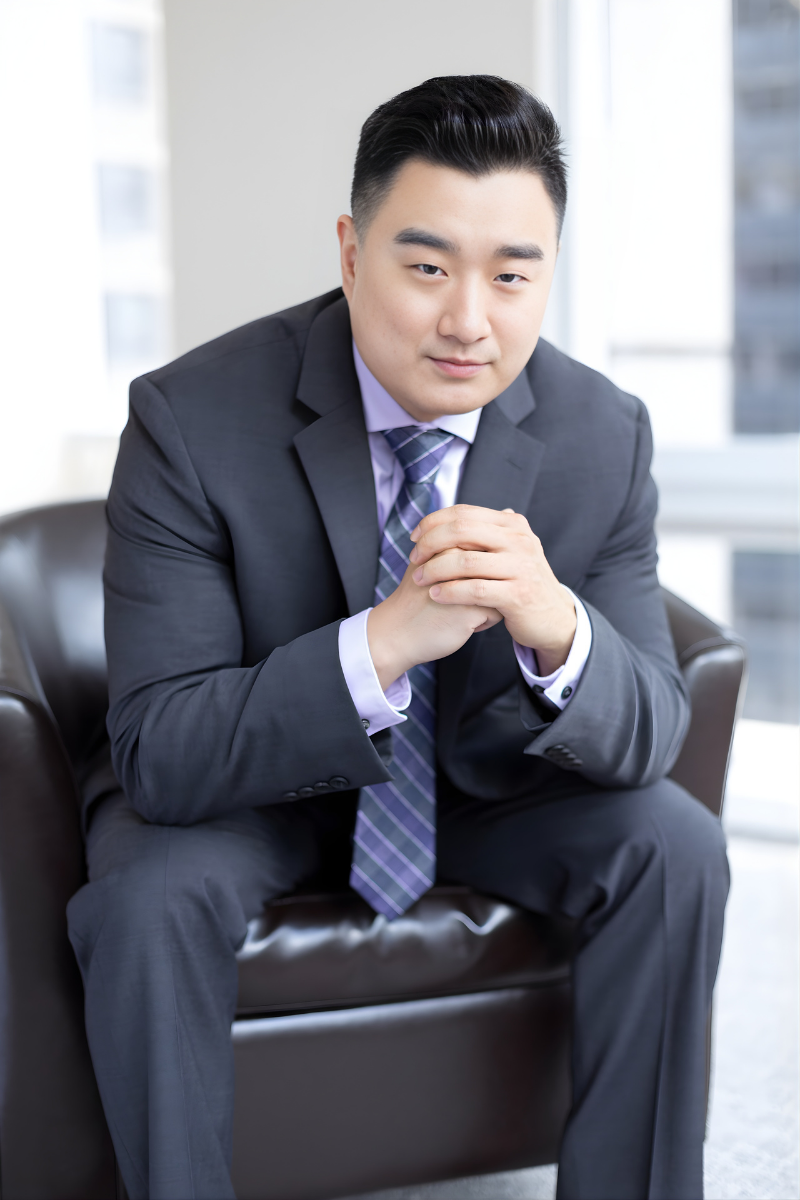 A young man with short black hair wearing a black T-shirt and blue jeans standing indoors against a neutral background.