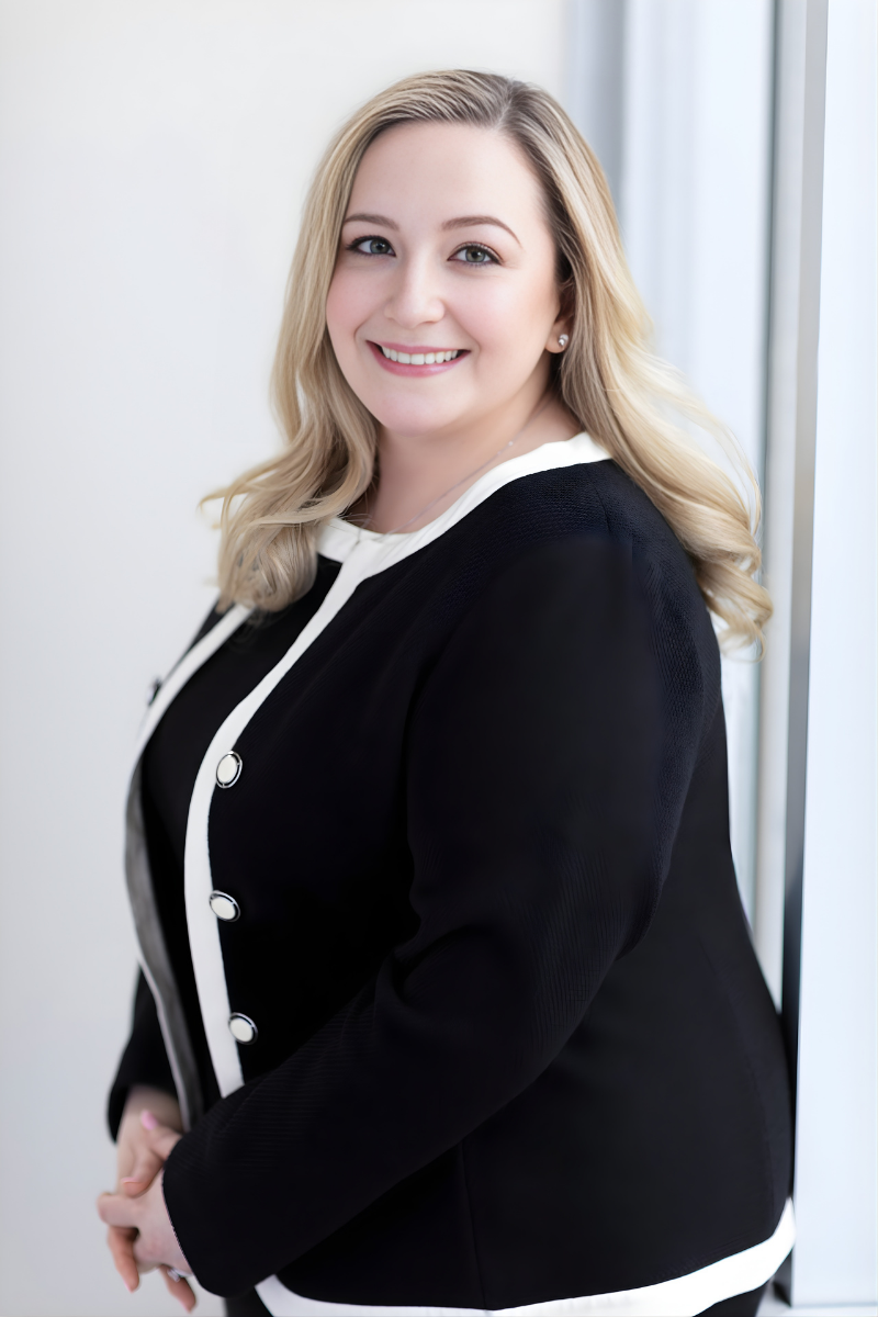 A smiling woman with blonde hair wearing a denim jacket and black dress, leaning on a gray chair in an indoor setting.