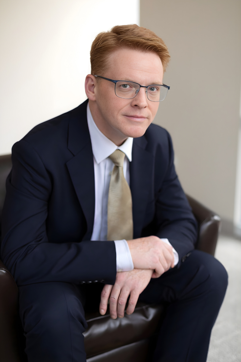 A man with red hair wearing a light gray shirt and a smartwatch sitting outdoors with green foliage in the background.