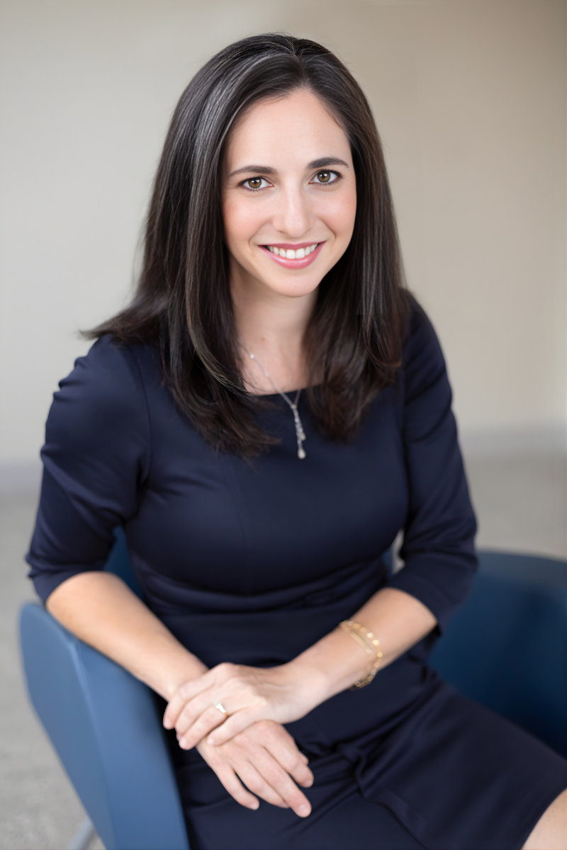 A woman sitting on a gray couch in a modern indoor space with green wall and wooden accents, wearing a white long-sleeve shirt and blue jeans, smiling at the camera.
