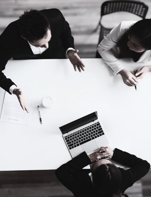 Three people having a discussion around a conference table with a laptop, a document, a pen, and a cup.