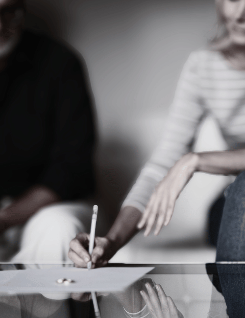 Two women are in a therapy session, one woman is writing on a notepad while the other sits nearby, with her hand on the table.