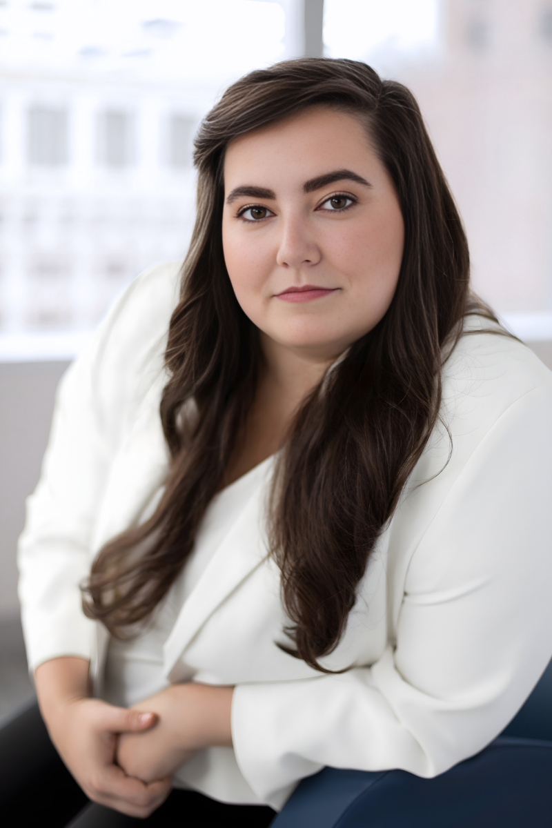 A woman with long brown hair and light skin is smiling at the camera, sitting indoors.